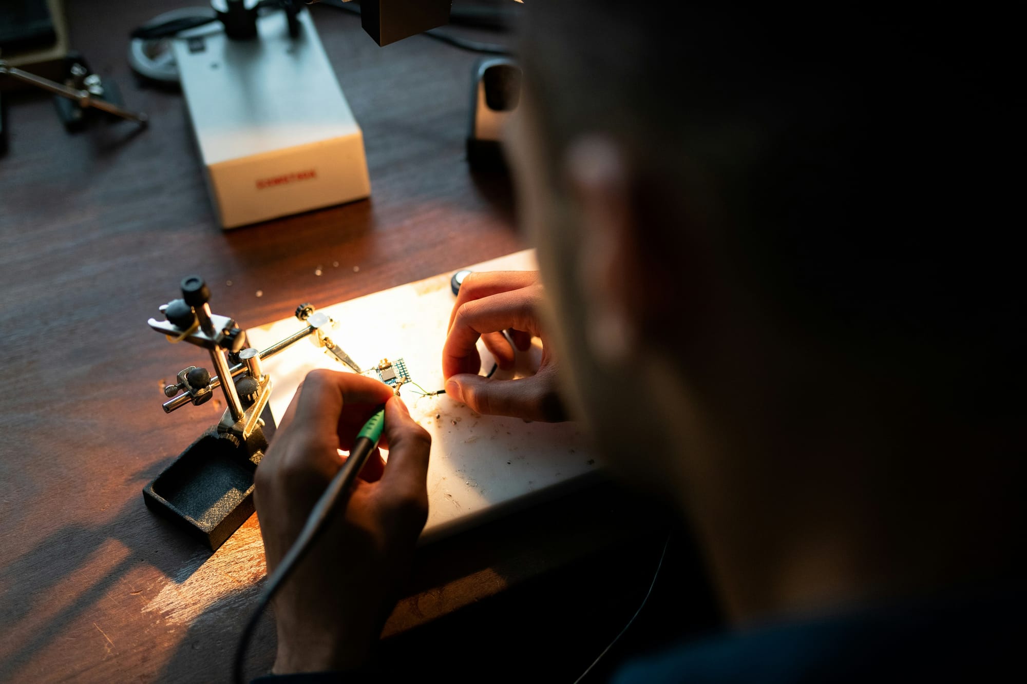 Mãos soldando componentes eletrônicos sob luz de bancada